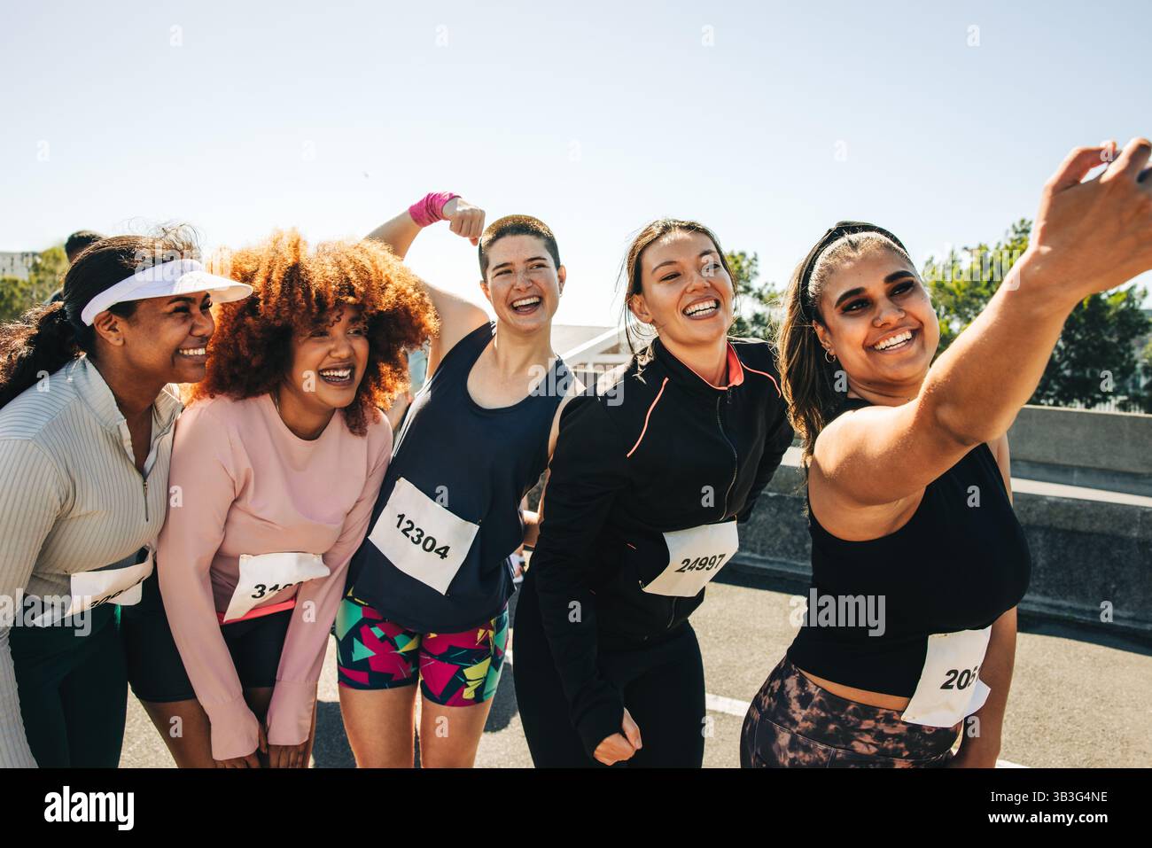 Un gruppo di donne vivace e diversificato sorride e scatta un selfie insieme durante una maratona. Indossano outfit sportivi e pettorali da running, show Foto Stock