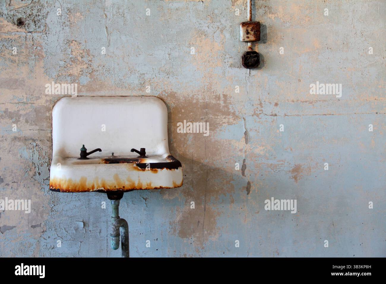 All'interno di una cella di prigione abbandonata sull'isola di Alcatraz, un vecchio lavandino arrugginito con rubinetti usurati si trova contro un muro blu-verde che si stacca. Foto Stock
