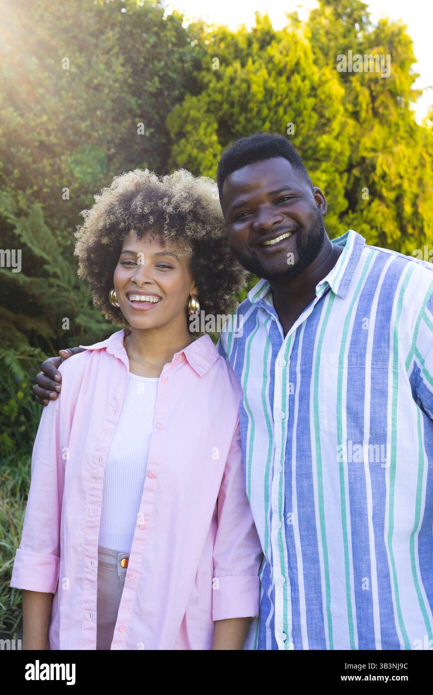 Coppia di persone sorridenti e diversificate in piedi vicini nel giardino sul retro, con luce solare delle lenti Foto Stock