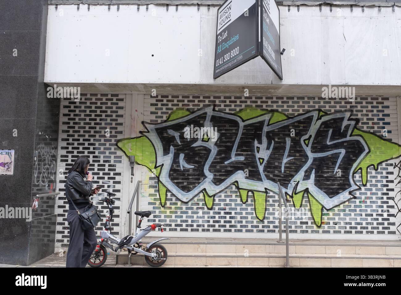 Woman with vape passa davanti a un negozio vuoto con graffiti in Sauchiehall Street, Glasgow, Scozia. Una bicicletta elettrica Niubility è collegata a un corrimano. Foto Stock