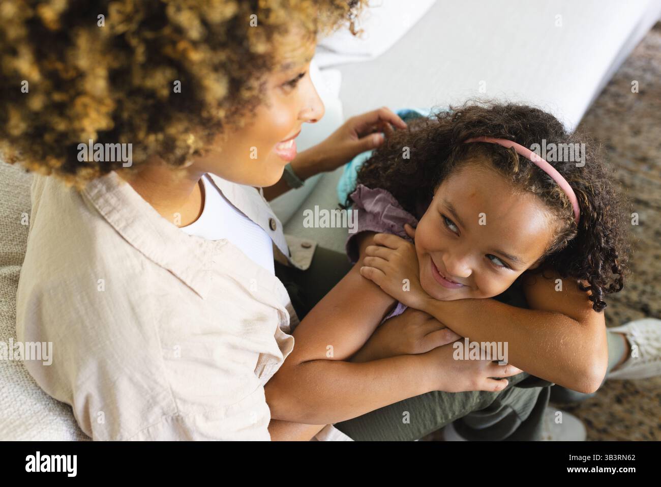 Sorridendo, madre e figlia diverse che si coccolano sul divano nel soggiorno, con cuscino e tappeto Foto Stock
