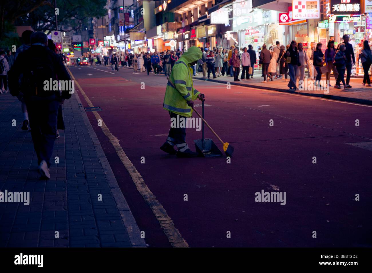 Il pulitore di strada con le attrezzature al neon spazza da solo in una serata vivace mentre la folla scorre accanto ai negozi luminosi Foto Stock