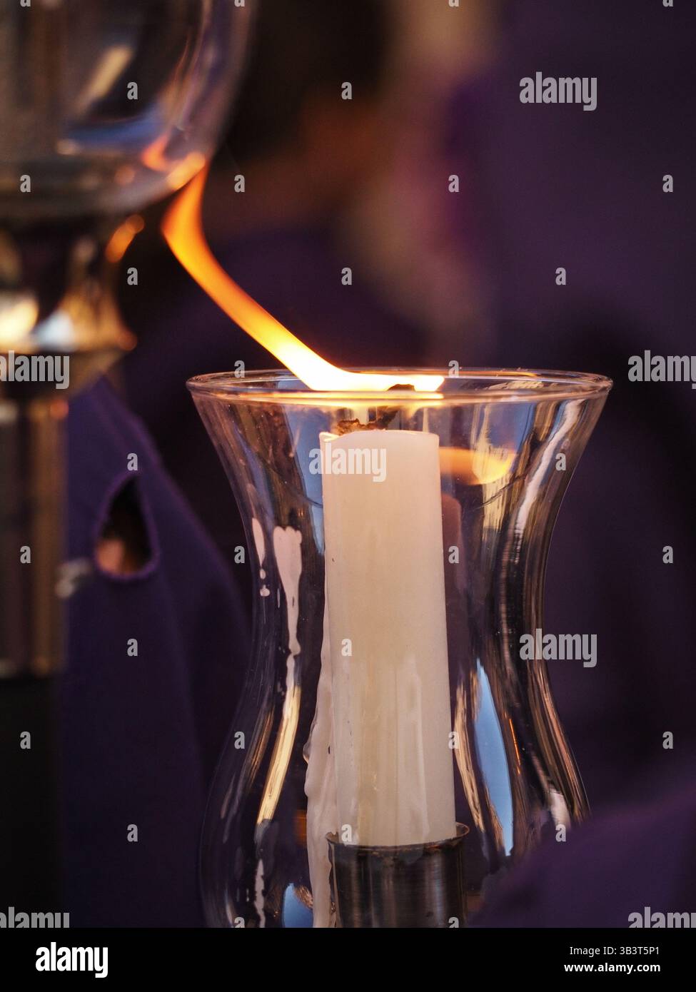 Una candela incandescente brucia all'interno di un supporto in vetro, emettendo una luce calda e serena. Sullo sfondo sfocato, candele e figure aggiuntive creano un morbido effetto Foto Stock