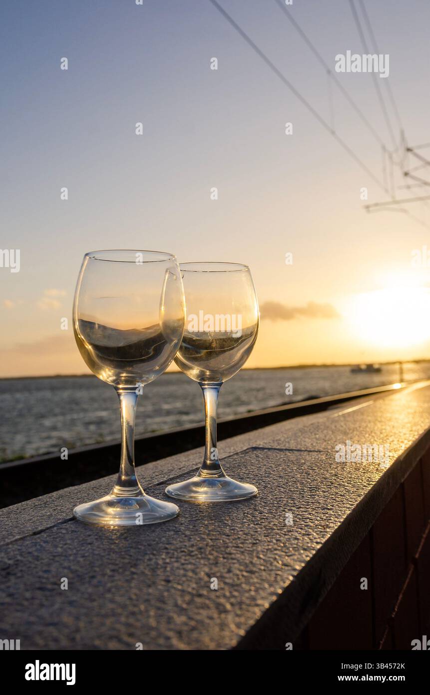 I bicchieri da vino vuoti si stagliano su una sporgenza che si affaccia sull'acqua tranquilla, riflettendo le calde sfumature di un pittoresco tramonto Foto Stock