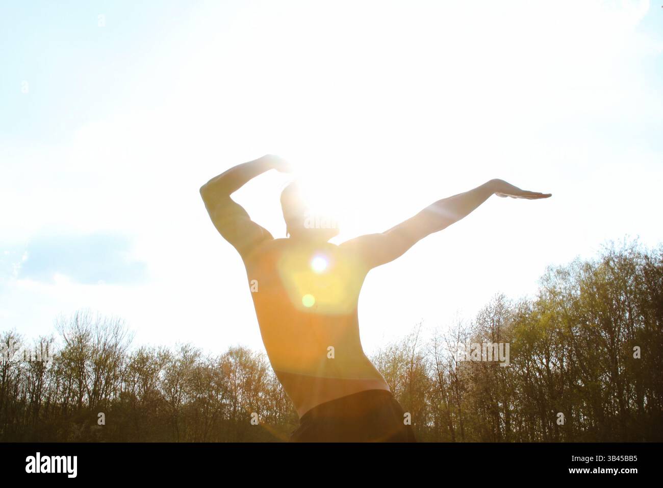 Uomo sportivo in posa contro la luce del sole con le mani in alto. Silhouette maschile che danzano in posa sportiva che evoca energia, buone vibrazioni, divertimento e divertimento. Foto Stock