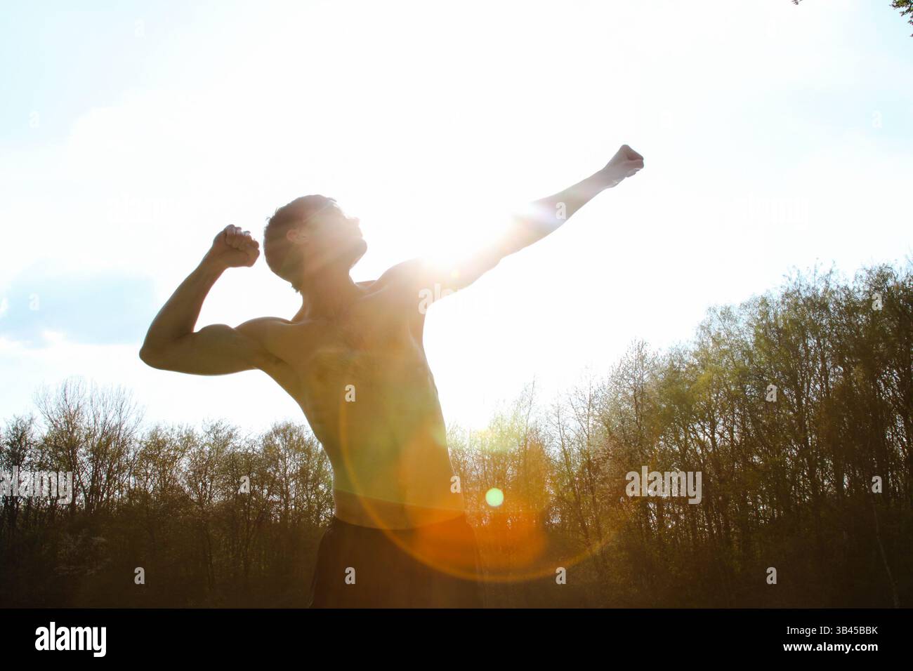 Uomo sportivo in posa contro la luce del sole con le mani in alto, uno stretto del braccio e un altro dietro la testa. Silhouette maschile in posa atletica. Foto Stock