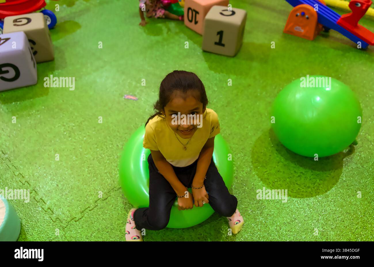 Un bambino allegro gioca su una vivace palla rimbalzante verde in un divertente parco giochi al coperto pieno di giocattoli. Foto Stock