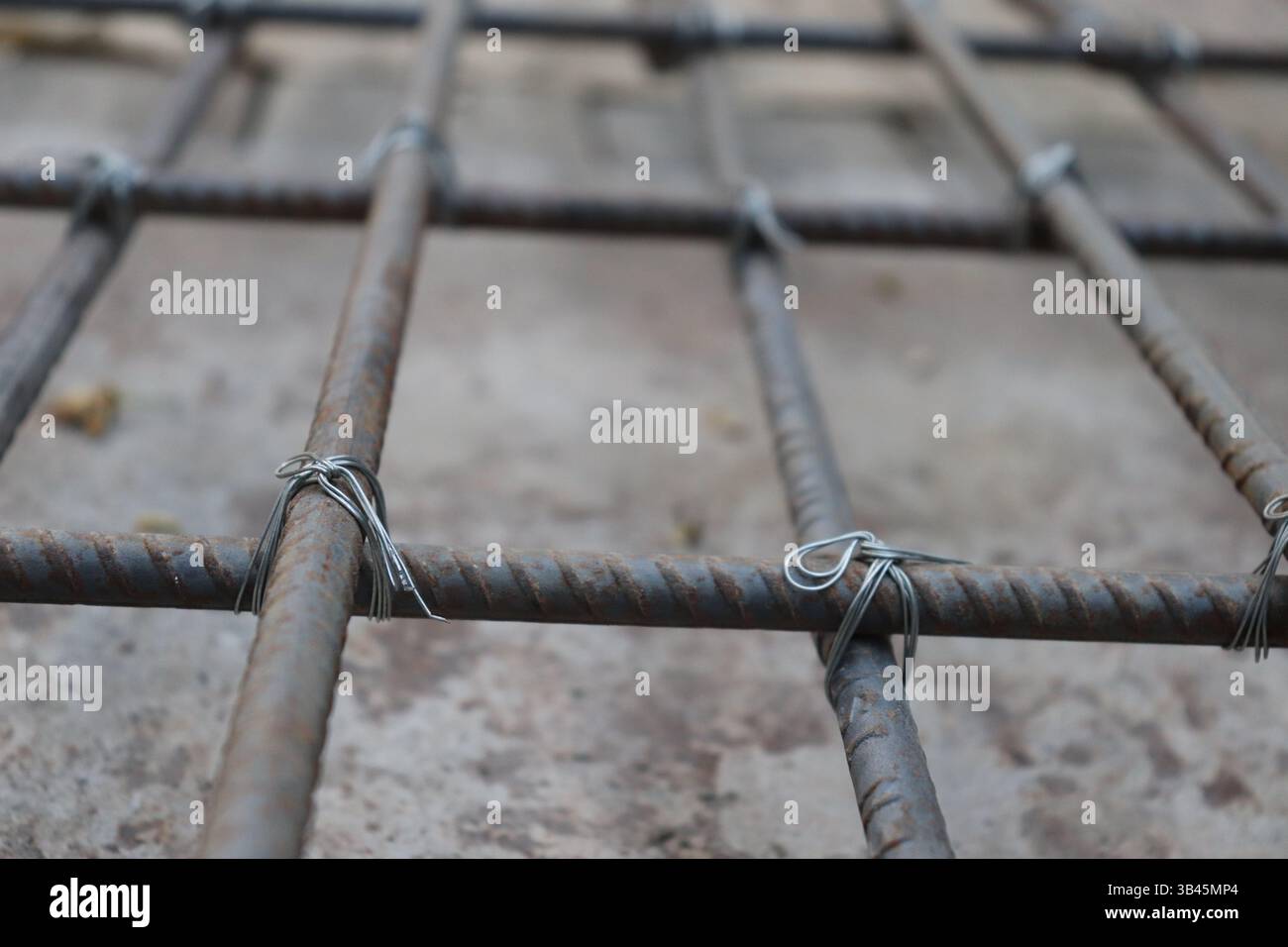 Primo piano mostra un tondino in metallo intrecciato legato insieme a un filo d'argento, formando una griglia su uno sfondo di cemento, evidenziando le basi della costruzione Foto Stock