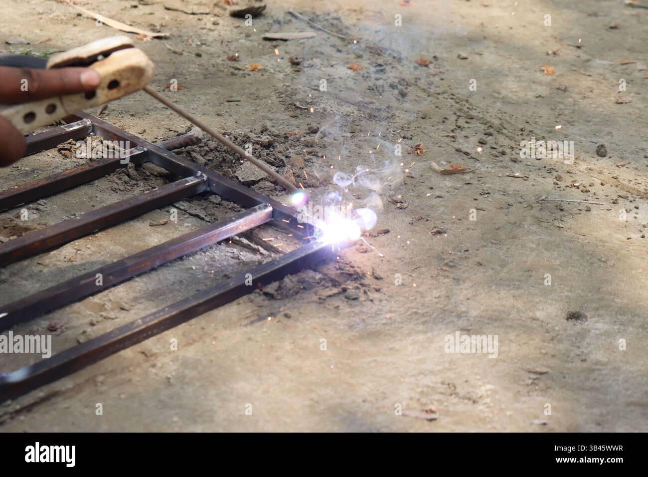 Persona dalla pelle scura che salda il telaio in metallo all'esterno con scintille luminose e fumo che creano interesse visivo, mettendo in mostra il lavoro industriale e la maestria artigianale. Foto Stock