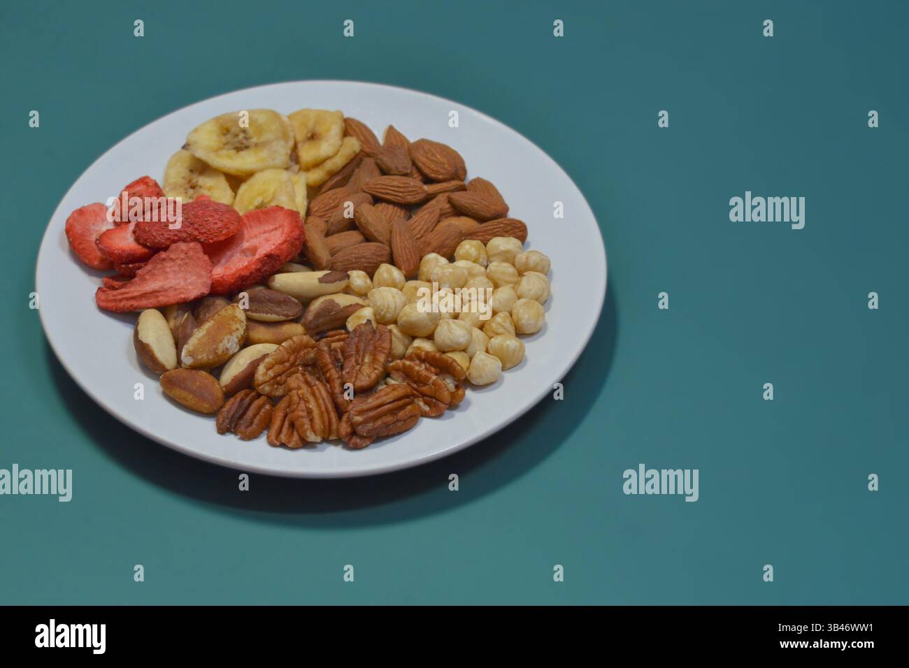 Una varietà di frutta secca e frutta secca su un piatto su sfondo blu. Nocciole, noci pecan, noci del brasile, mandorle, fragole secche e banane. Il concetto Foto Stock