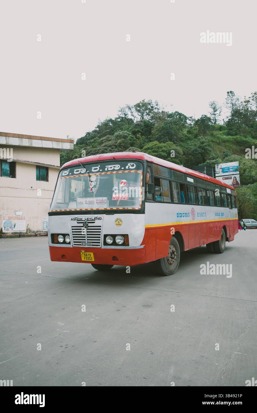 Un autobus rosso e bianco KSRTC Ashok Leyland è parcheggiato su una strada in una zona collinare, con vegetazione ed edifici sullo sfondo sotto un cielo coperto. Foto Stock