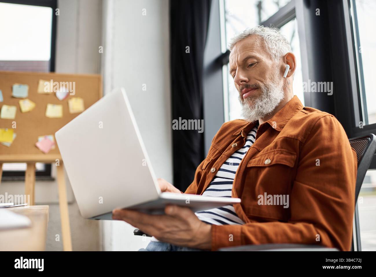 Un uomo anziano in abbigliamento casual lavora diligentemente su un notebook in un ufficio organizzato. Foto Stock
