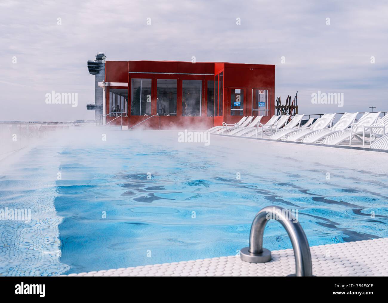 Moderna piscina sul tetto presso il TWA Hotel, JFK Airport a New York City, con vapore in aumento, lettini bianchi, un audace bar rosso e la torre di controllo. Foto Stock
