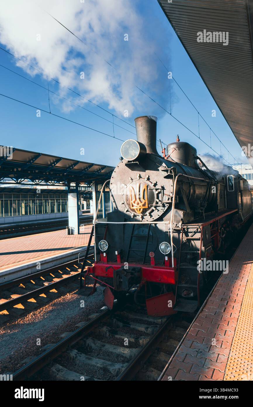 Storico treno a vapore in piedi su una piattaforma della stazione sotto un cielo blu. Emissione di vapore, che rappresenta il trasporto e la storia d'epoca Foto Stock