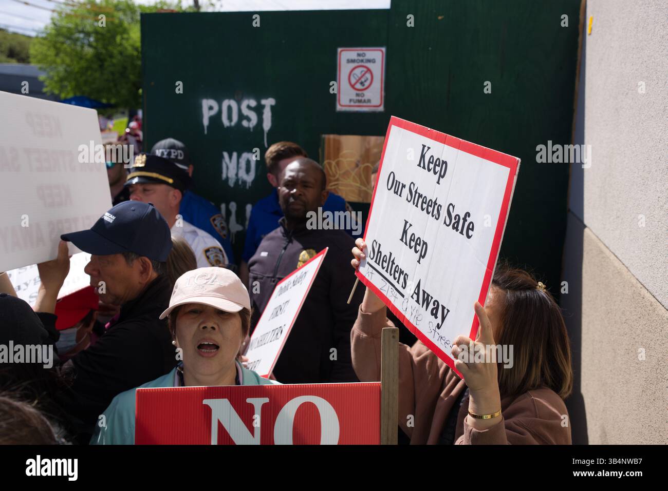 Si apre una scena di protesta, con persone che tengono in mano cartelli che recano "Keep our Streets Safe, Keep Shelters Away" e "NO". Lo sfondo include un Foto Stock
