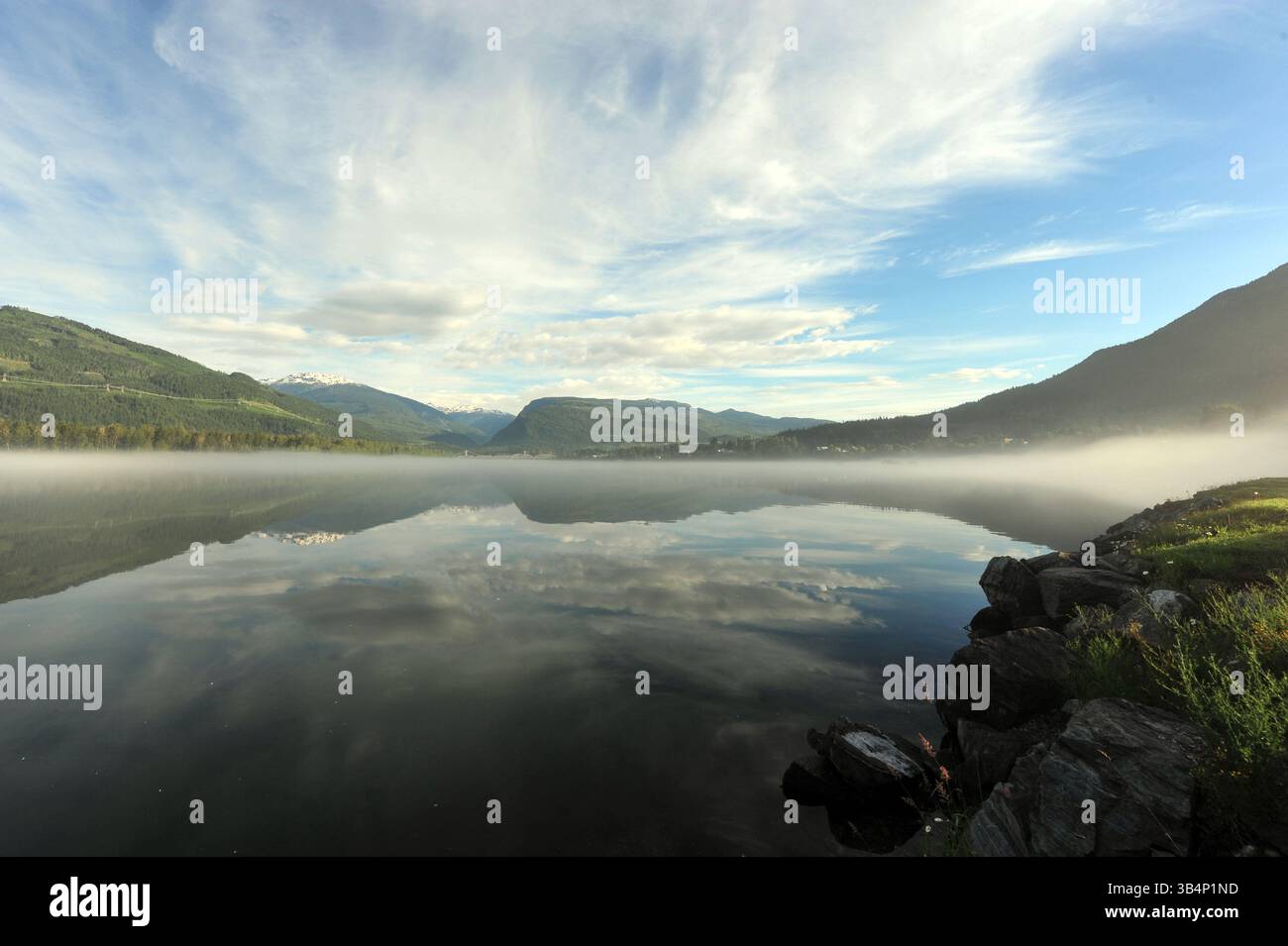 Un lago di montagna ferma riflette morbide nuvole mattutine e pendii boscosi mentre la nebbia permane vicino al litorale in una tranquilla giornata estiva. Foto Stock
