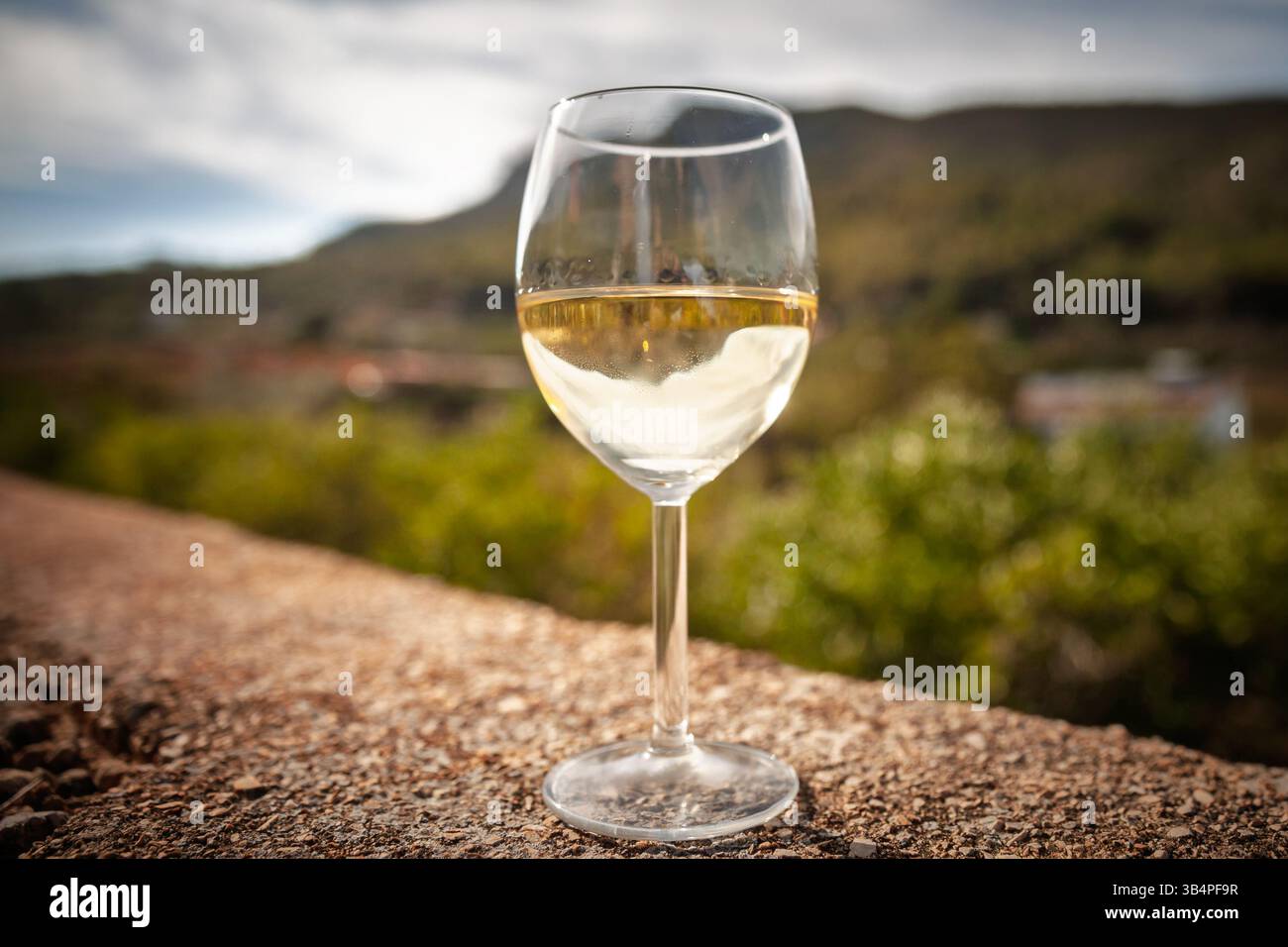 Un buon bicchiere di vino bianco spagnolo ghiacciato si erge su una terrazza in pietra; le colline e i villaggi mediterranei sfocati creano un'atmosfera rilassata sotto la p Foto Stock