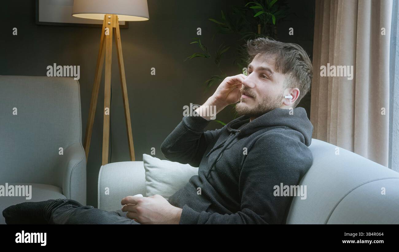 Giovane che ascolta musica con gli auricolari wireless mentre si rilassa a casa su un divano Foto Stock
