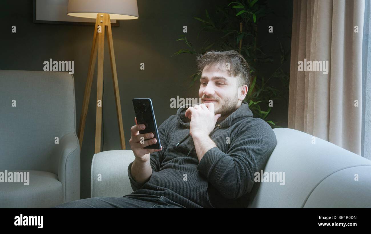 Giovane uomo che si rilassa su un divano e sorride mentre utilizza lo smartphone a casa in un accogliente salotto Foto Stock