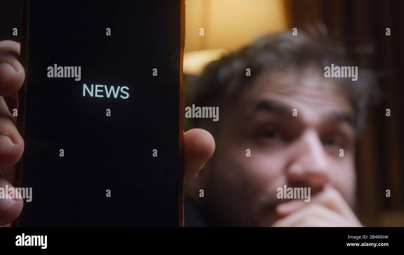 Giovane eccitato che punta sullo schermo dello smartphone che visualizza le NOTIZIE, reagisce entusiasticamente alle ultime notizie in un ambiente interno accogliente con Foto Stock