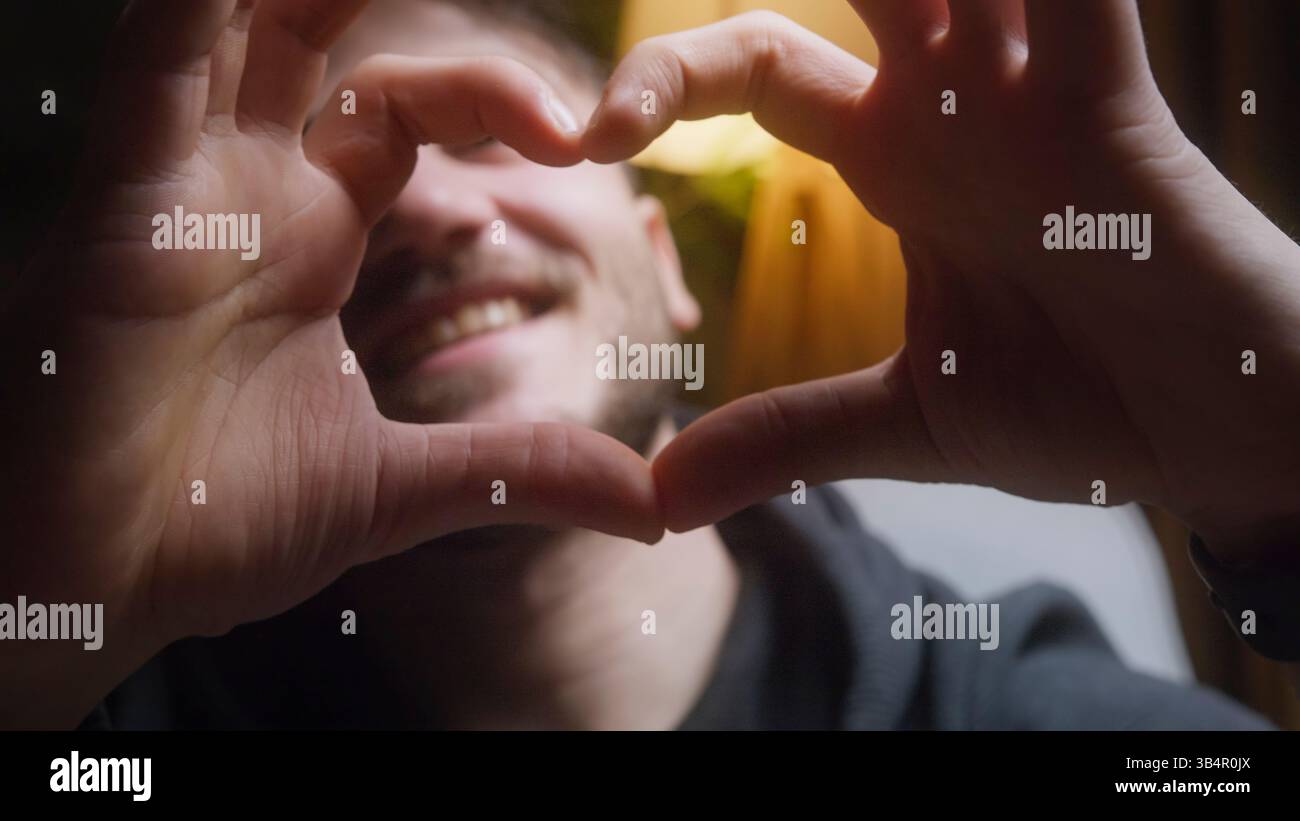 Un giovane sorridente che crea una forma a cuore con le mani per esprimere amore e positività in casa Foto Stock