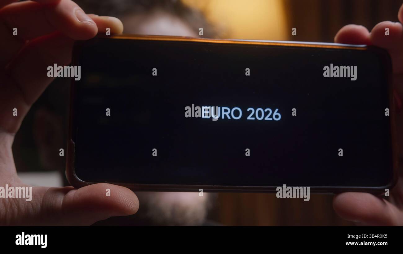 Giovane in possesso di smartphone che visualizza testo EURO 2026 sullo schermo nella stanza buia Foto Stock