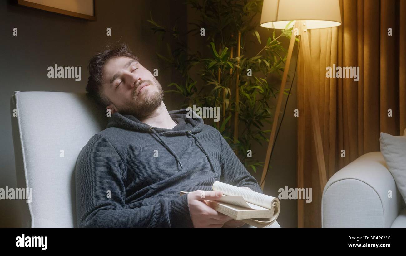 Young Man dorme tranquillamente in poltrona dopo aver letto un libro di notte Foto Stock