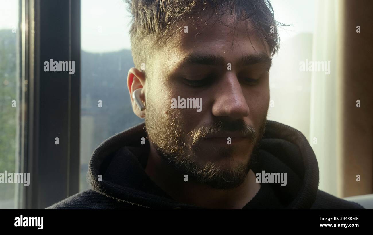 Giovane uomo che si rilassa con gli occhi chiusi mentre ascolta la musica sugli auricolari vicino alla finestra Foto Stock