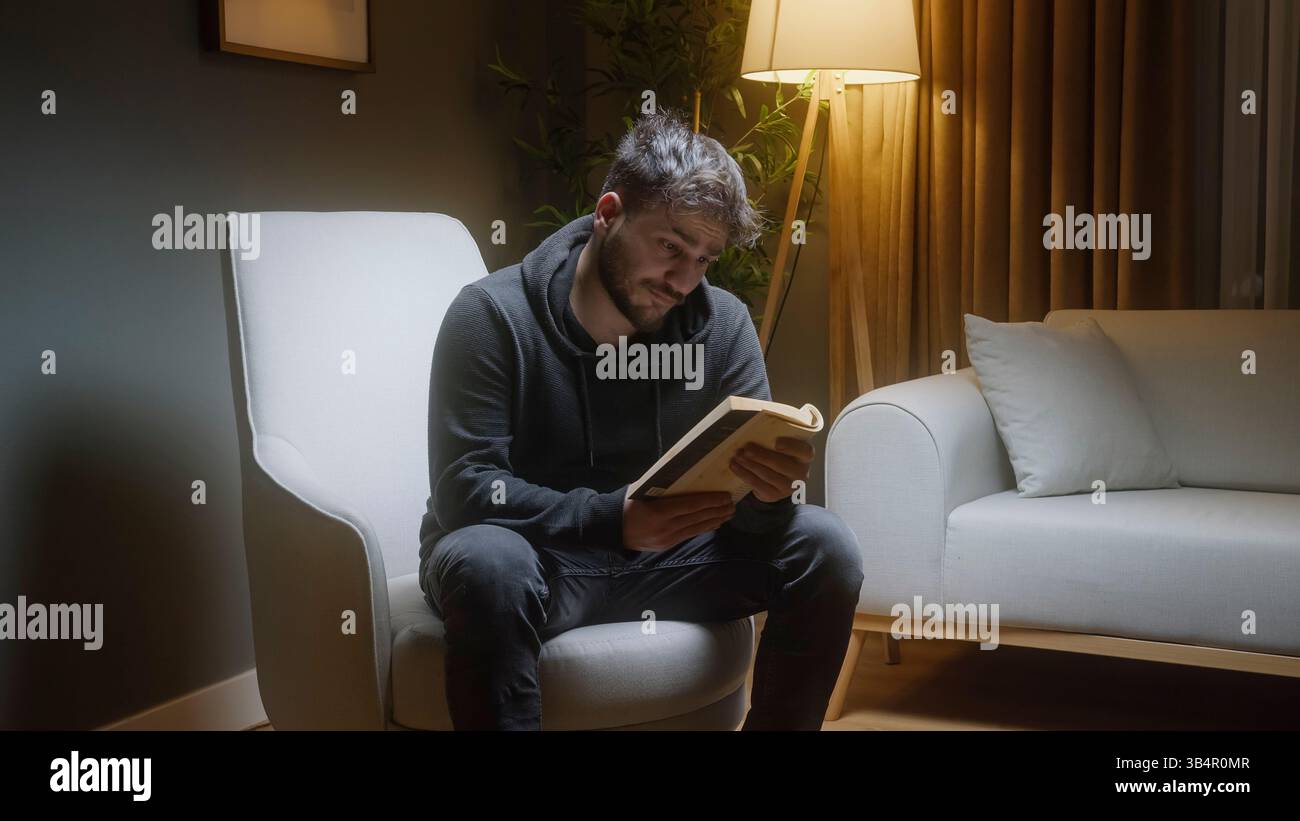 Giovane uomo che si rilassa a casa leggendo un libro in un'illuminazione serale accogliente Foto Stock