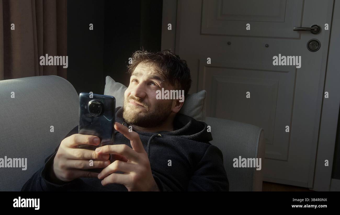 Uomo che si rilassa su un divano mentre sfoglia il suo smartphone in luce naturale Foto Stock