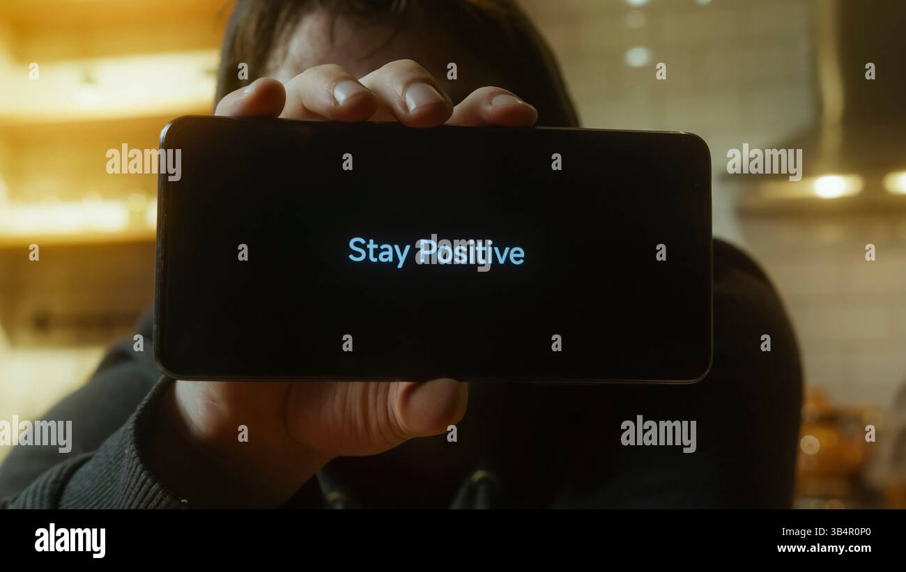 Uomo sorridente che tiene in mano lo smartphone che visualizza il messaggio resta positivo in Warm Indoor Lighting Foto Stock