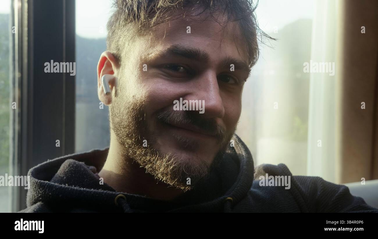 Giovane sorridente mentre ascolta la musica e guarda la fotocamera in luce naturale Foto Stock