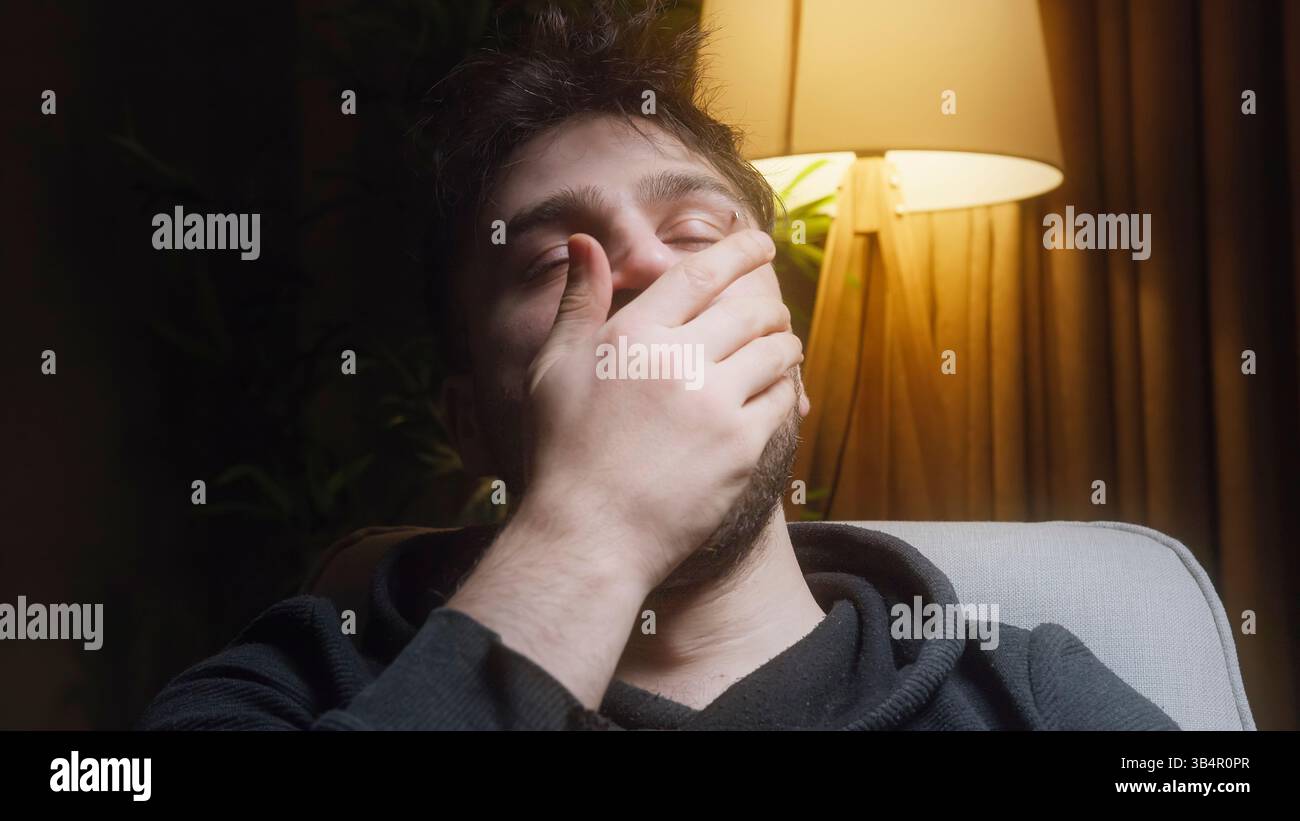 Giovane seduto su un divano in una stanza poco illuminata, coprendo il viso con la mano in un gesto di disagio emotivo, stanchezza o frustrazione Foto Stock