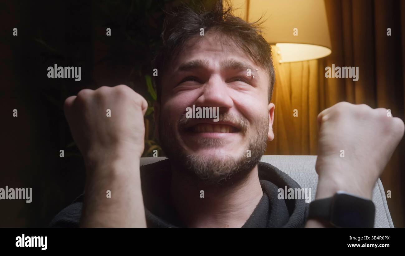 Giovane eccitato che tira con i pugni alzati mentre celebra il successo al chiuso di notte Foto Stock