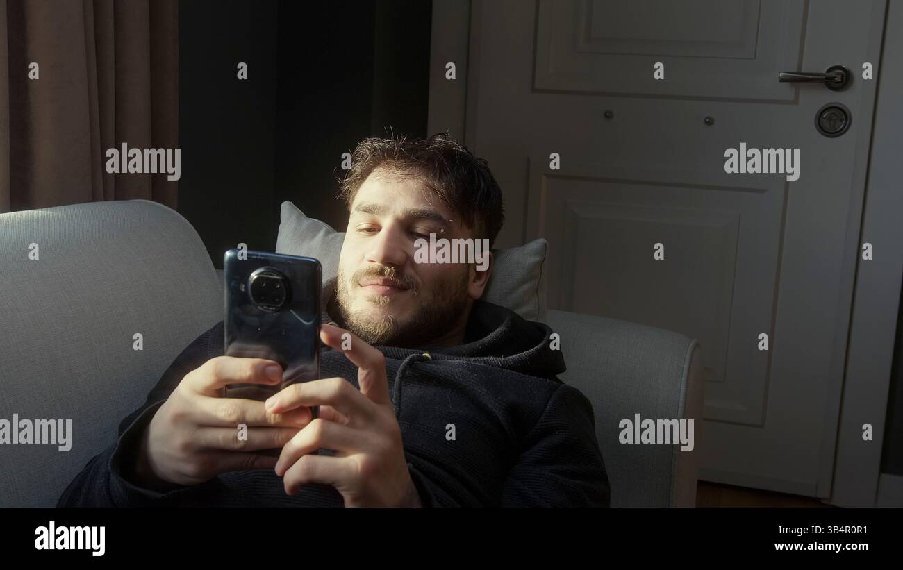 Giovane uomo che si rilassa sul divano mentre sfoglia il suo smartphone in luce naturale Foto Stock