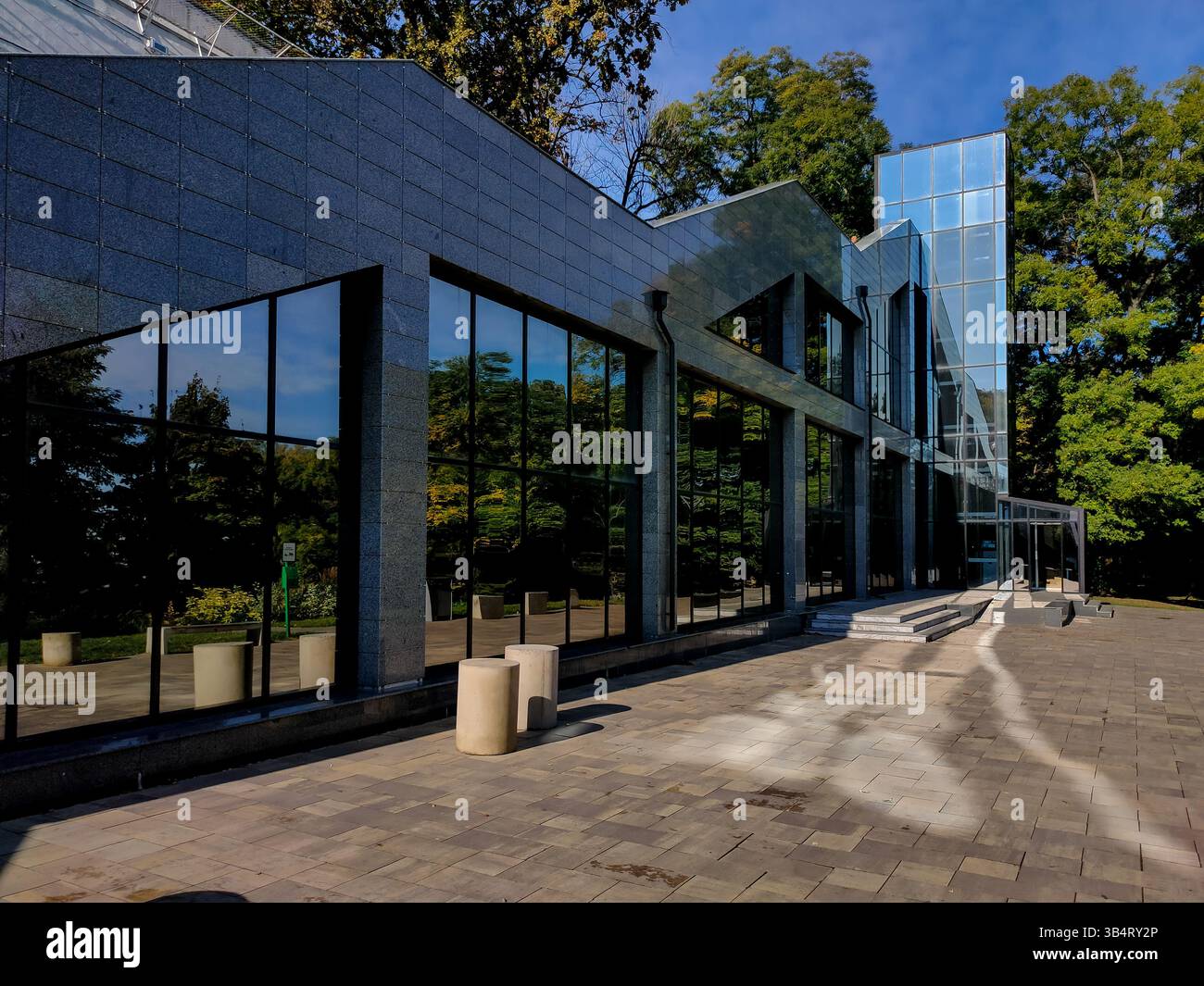 Struttura contemporanea con grandi finestre riflettenti e un'elegante facciata in granito, circondata da vegetazione lussureggiante e cielo blu limpido, a creare un luminoso e Foto Stock