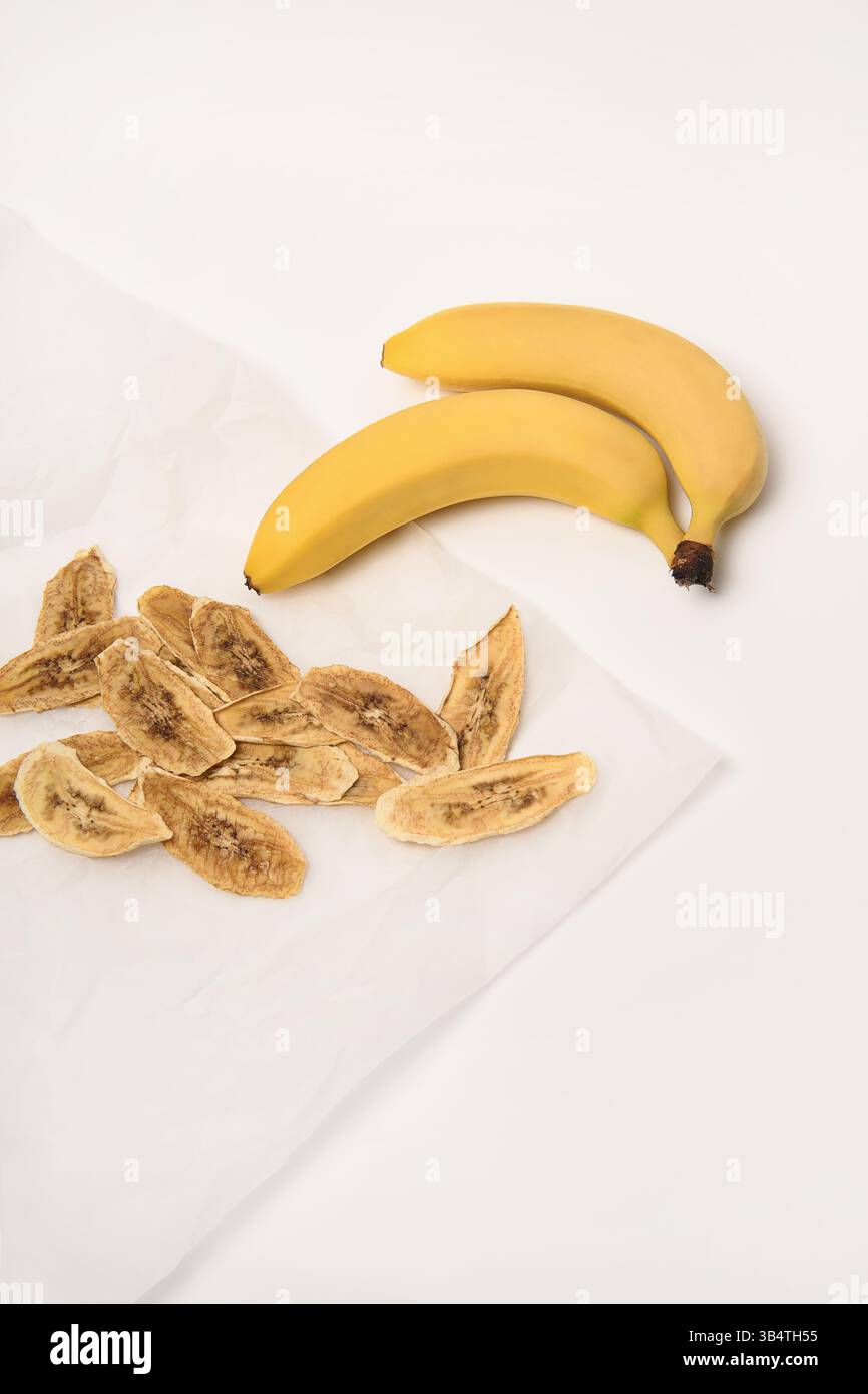 Sfondo bianco. Due banane fresche, accanto alle quali si trovano fette di banana essiccate su carta pergamena. Foto Stock