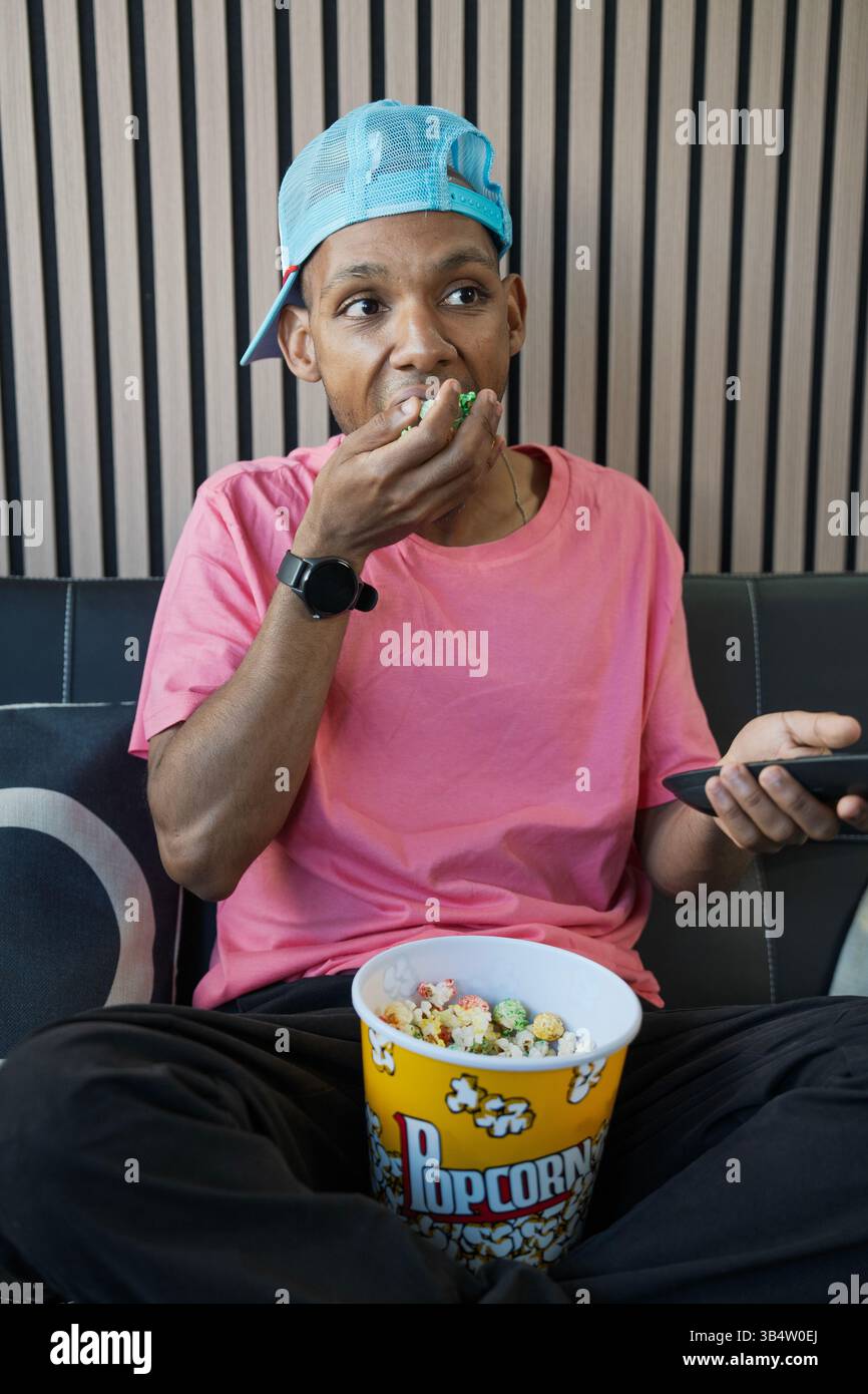 Giovane seduto sul divano, mangiando popcorn dal secchio e tenendo il telecomando, godendo dell'intrattenimento domestico Foto Stock