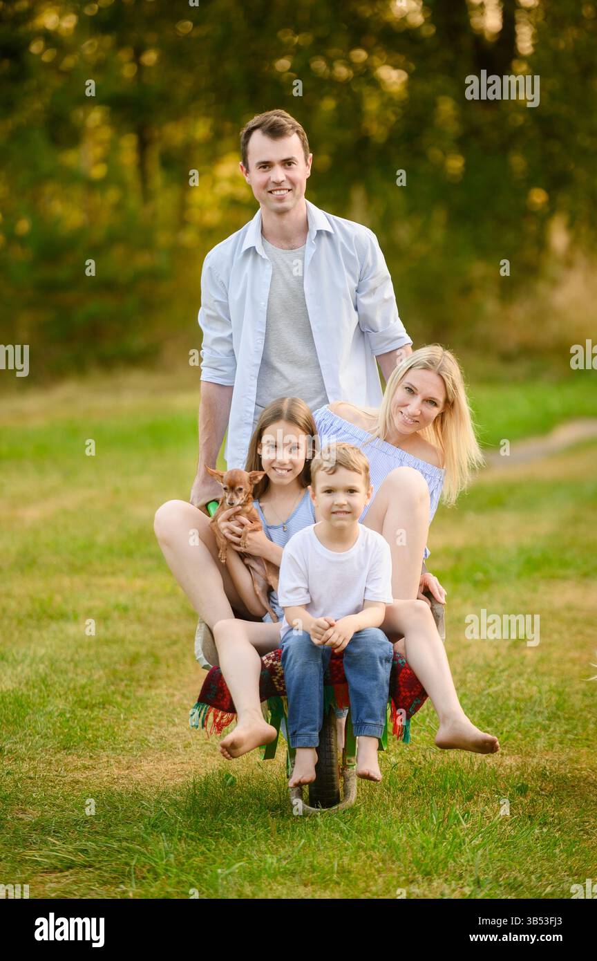 Famiglie che si rilassano in campagna e si divertono. Uomo che spinge sua moglie, suo figlio e sua figlia in carriola da giardino, con il cane. Concetto di giardinaggio, famiglia di campagna Foto Stock