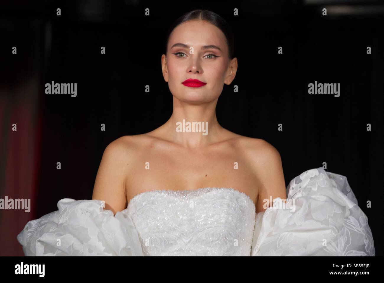 Primo piano di una donna con i capelli scuri tirati indietro, che indossa un abito da sposa senza spalline con perline bianche e maniche soffici. Ha il rossetto rosso e il sottile ey Foto Stock