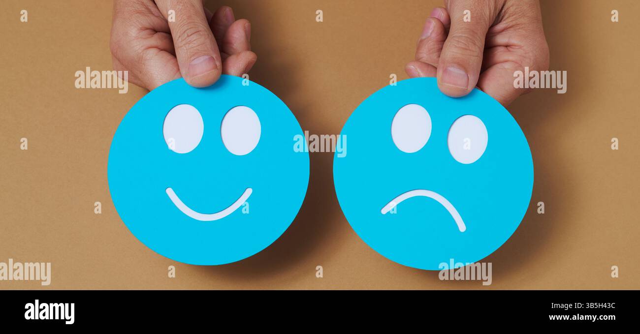 un primo piano delle mani di un uomo che ha un volto felice di carta blu, nella mano sinistra, e un volto triste di carta blu, nella mano destra, su uno sfondo marrone Foto Stock