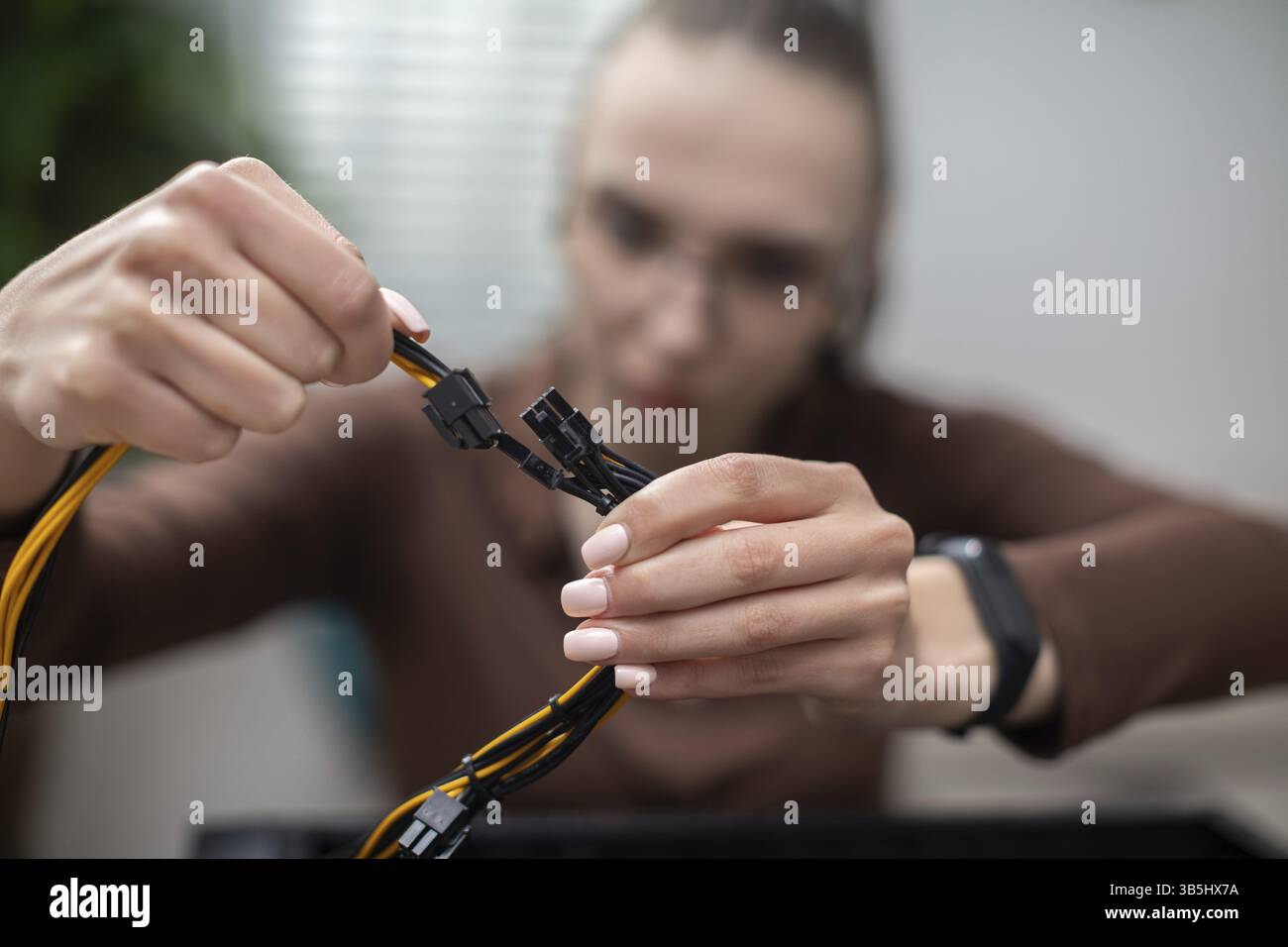 Primo piano delle mani di una donna che tiene dei cavi gialli. Sullo sfondo c'è una figura sfocata di una donna che indossa gli occhiali e che viene pettinata in una coda di cavallo. La Wom Foto Stock