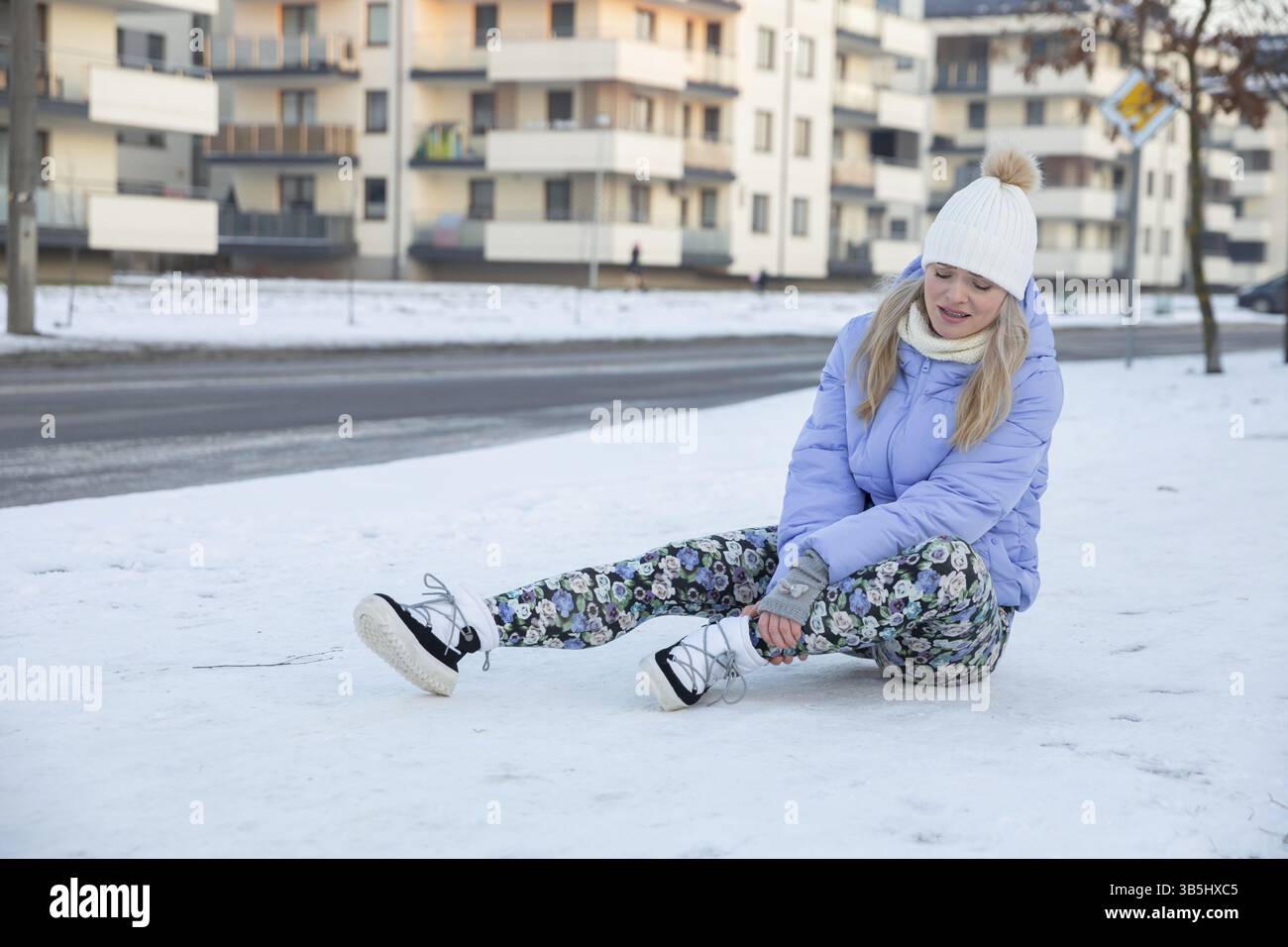 Un giovane imprudente scivolò sulla neve e cadde. Si siede e massaggia la caviglia dolorosa. Accanto a lei c'è la strada. Dall'altra parte della strada, c Foto Stock