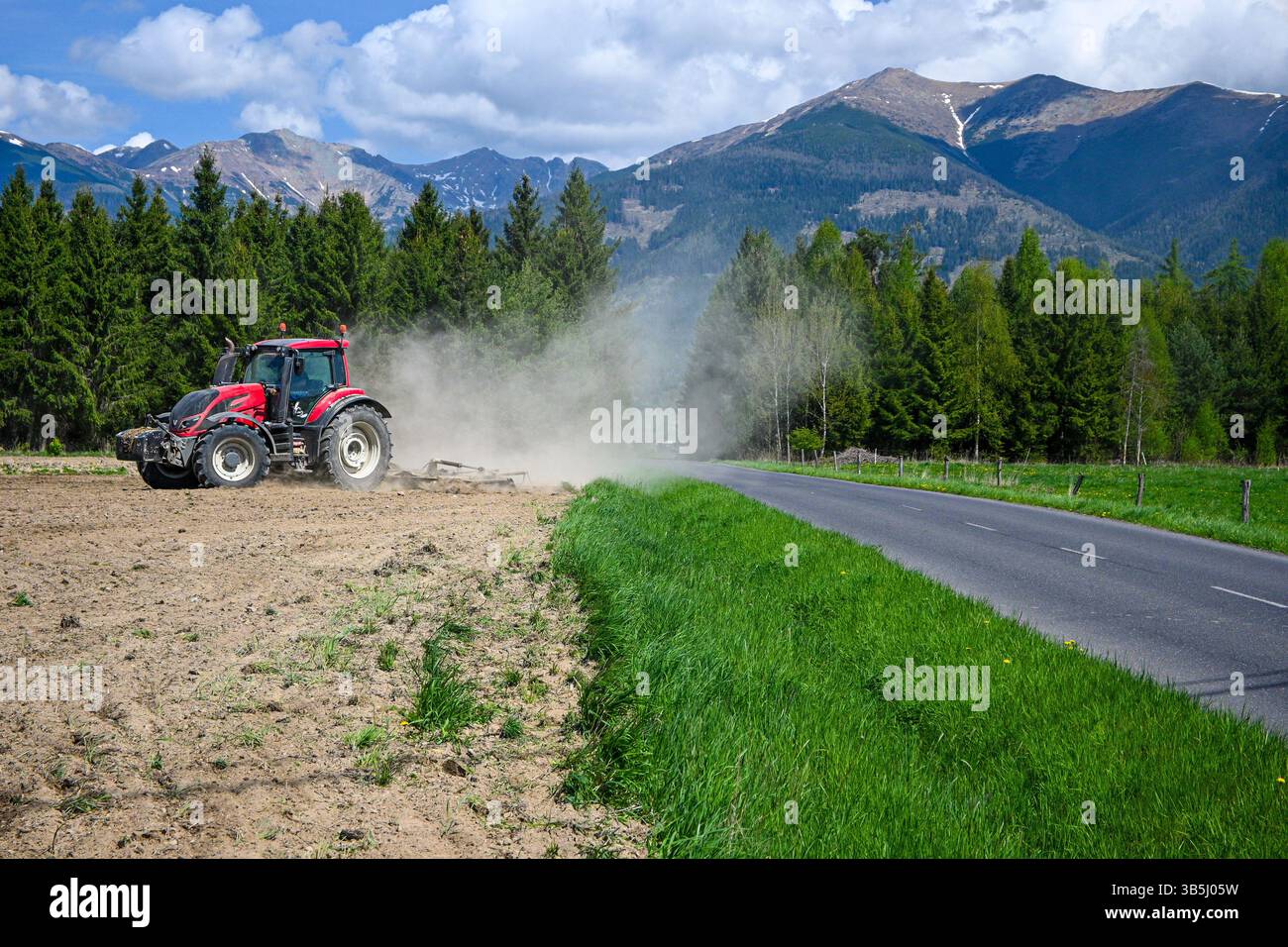 Trattore rosso aratura di un campo agricolo vicino a una strada asfaltata con boschi e montagne sullo sfondo nelle giornate di sole. Foto Stock