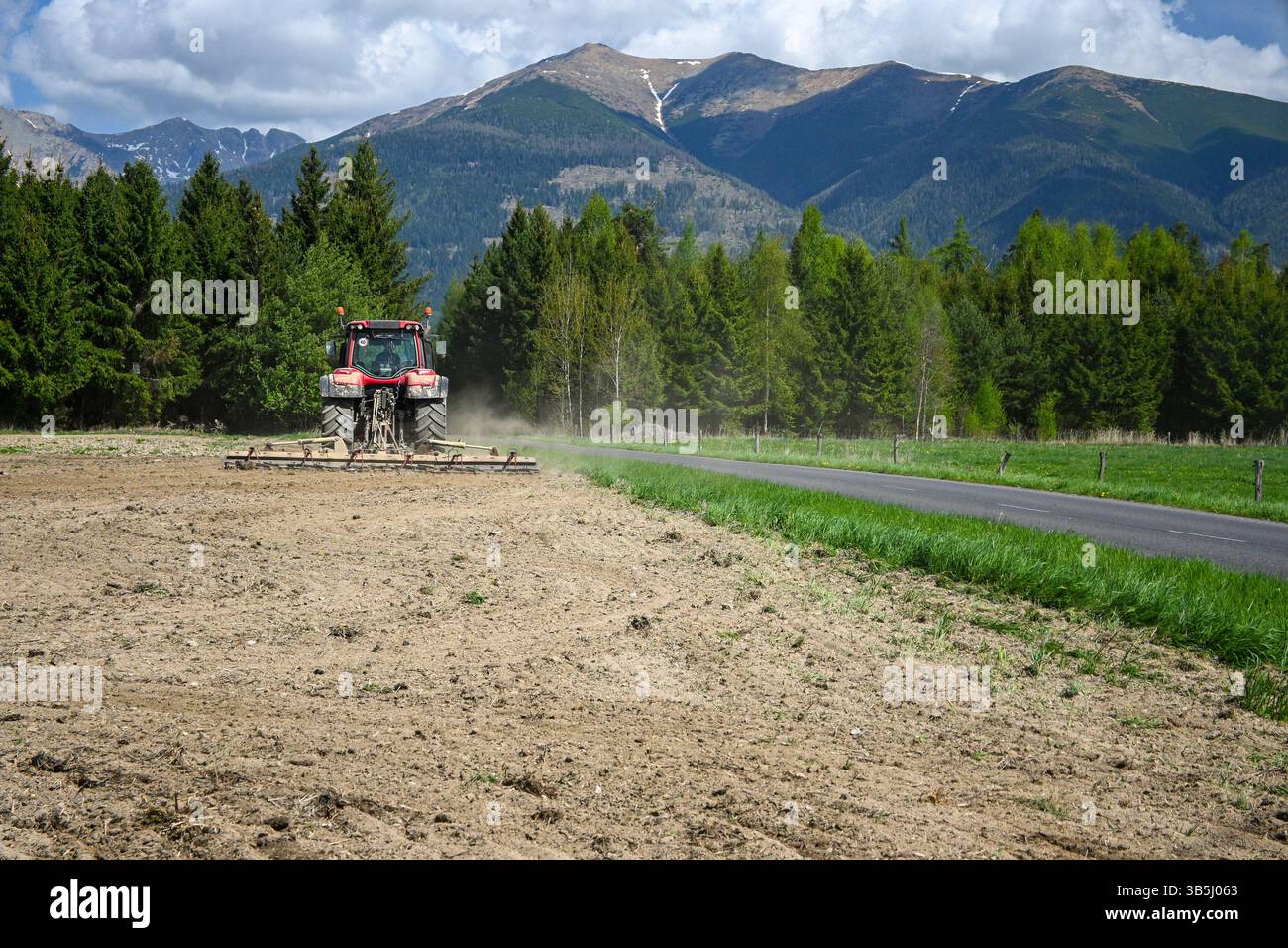 Trattore rosso aratura di un campo agricolo vicino a una strada asfaltata con boschi e montagne sullo sfondo nelle giornate di sole. Foto Stock