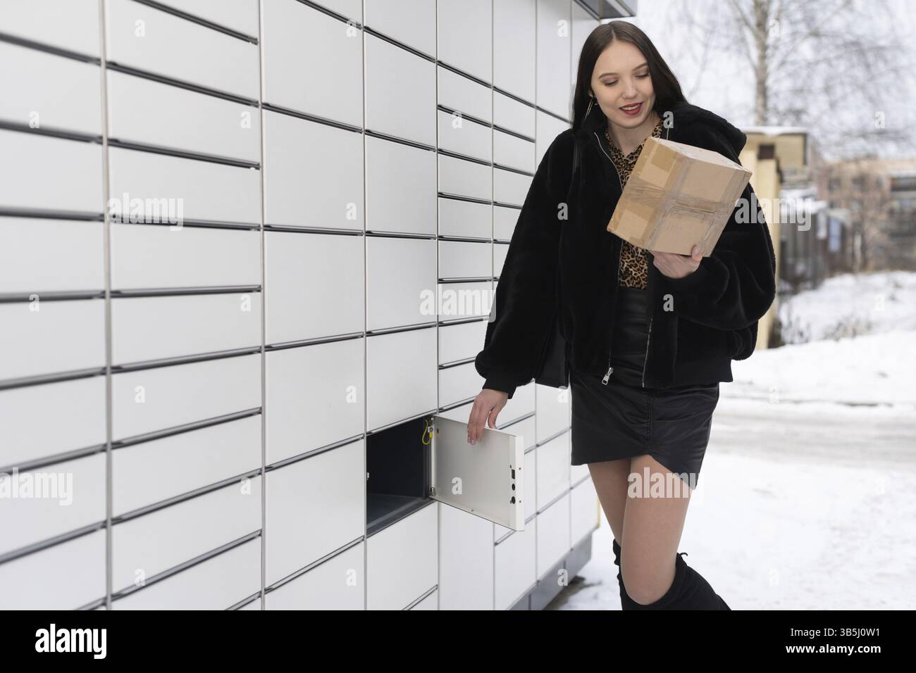 In inverno uno studente attraente raccoglie e invia un pacco tramite un armadietto. Macchina automatica per l'invio e la ricezione di lettere e pacchi Foto Stock