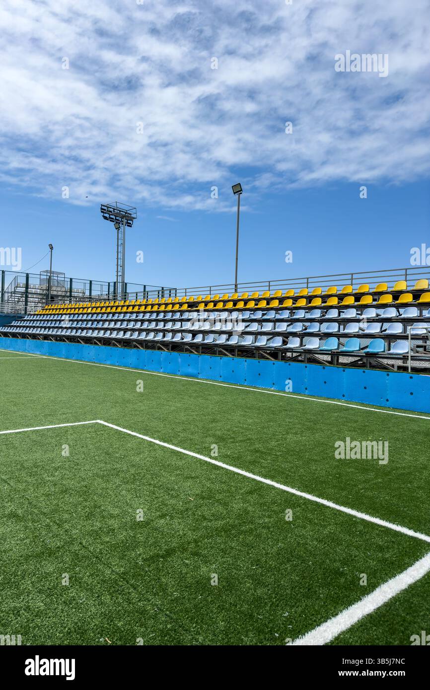 Vista verticale delle tribune vuote dello stadio con sedili colorati e campo da calcio artificiale in erba sotto il cielo blu. Struttura sportiva all'aperto, non ammessa la presenza di persone. Foto Stock