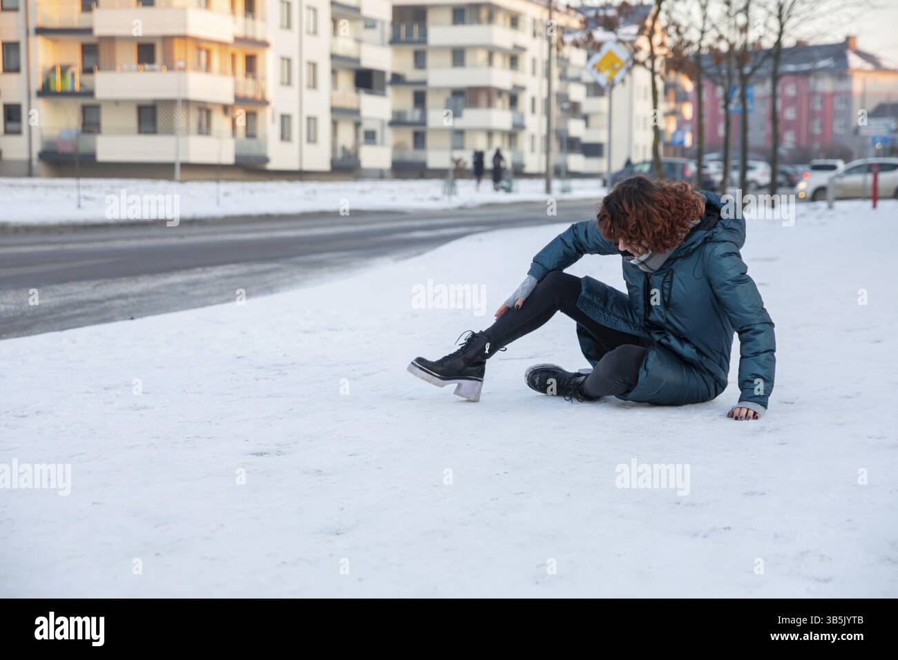 Una ragazza ha perso l'equilibrio camminando su un marciapiede della città senza neve. Si siede sulla neve e cerca di alzarsi. Accanto a lei c'è una strada trafficata. Attraverso il stre Foto Stock