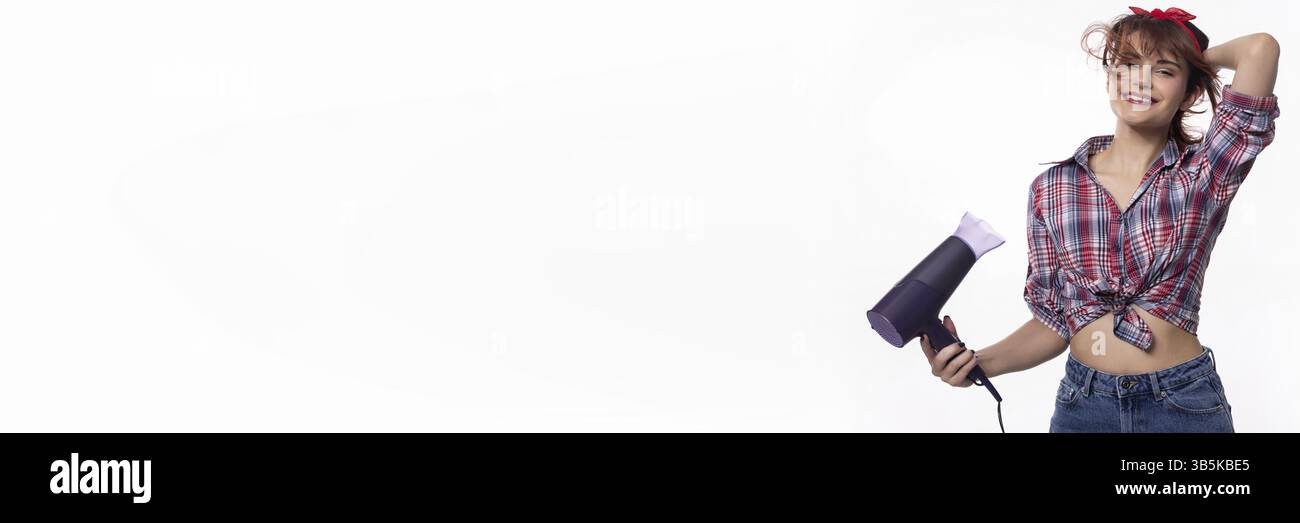 Una giovane donna elegante e alla moda regge giocosamente un asciugacapelli mentre mostra il suo look divertente ed elegante Foto Stock