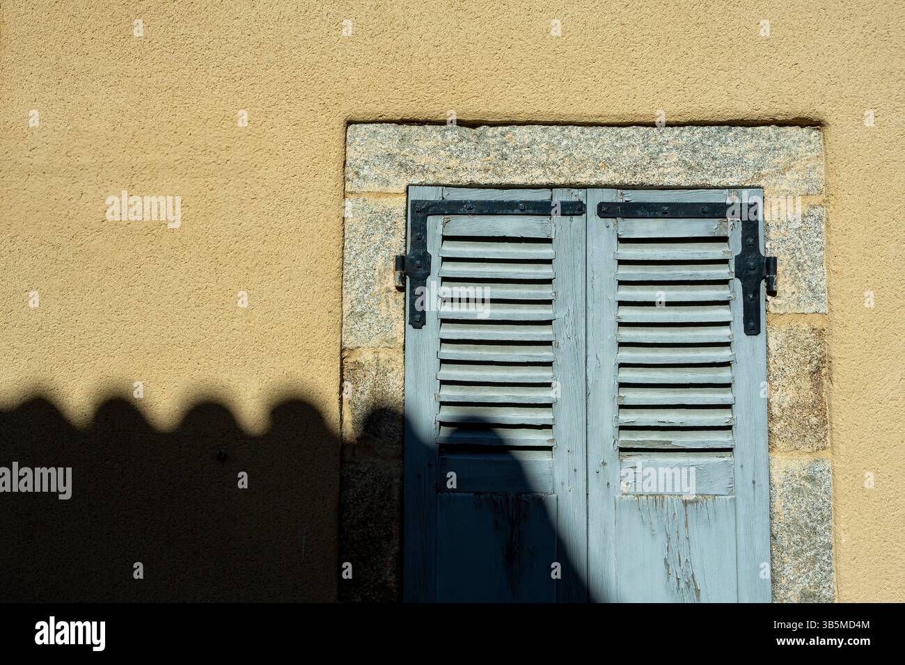 Un paio di classiche persiane in legno in una morbida tonalità blu si adagia su una parete marrone chiaro. La cornice in pietra aggiunge carattere alle ombre Foto Stock